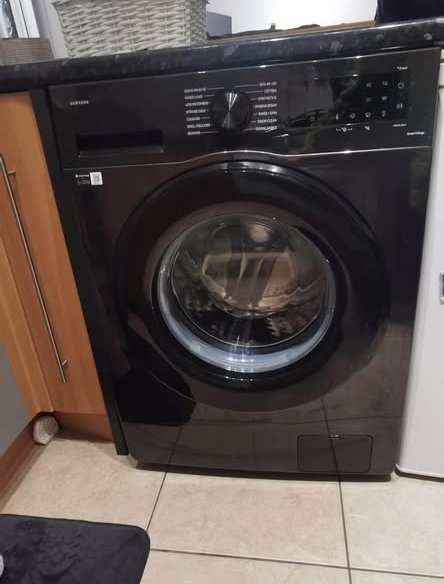 Black Samsung 12 kg (wash) 7 kg (dry) WD12FG5B34BBTL front‑load washer dryer installed under a countertop, showing its round glass door and top control panel